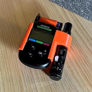 May include: An orange 3D-printed holder containing a black OneTouch Select Plus Flex blood glucose meter and a black lancing device. The meter has a digital display and several buttons. The lancing device has a numbered dial.