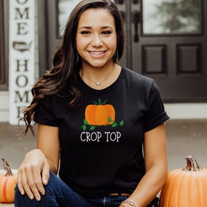 May include: A black t-shirt with a white pumpkin graphic and the text "Crop Top". The model is wearing blue jeans and sitting on a wooden step.