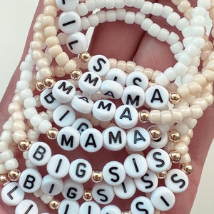 May include: A set of white beaded bracelets with gold beads and black letter beads spelling out "Lil Sis", "Big Sis", and "Mama".