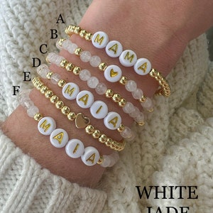 May include: Six white beaded bracelets with gold beads and gold letter charms spelling out the names "MAIA" and "MAMA".  The bracelets are stacked on a person's wrist.  The text "WHITE JADE" is written below the bracelets.