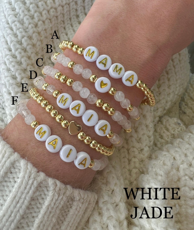 May include: Six white beaded bracelets with gold beads and gold letter charms spelling out the names "MAIA" and "MAMA".  The bracelets are stacked on a person's wrist.  The text "WHITE JADE" is written below the bracelets.
