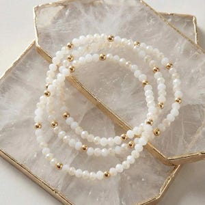 May include: Three white beaded bracelets with gold accents are displayed on a white and gold tray. The bracelets feature small, faceted beads and larger gold beads. The tray has a gold border.