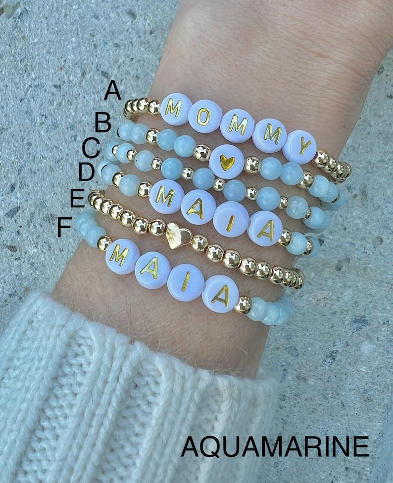 May include: Six beaded bracelets with gold beads and white beads with gold lettering. The bracelets spell out "MOMMY" and "MAIA". The beads are aquamarine.