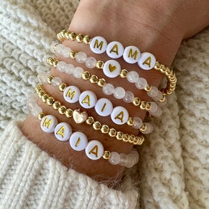 Peut inclure: Un ensemble de bracelets en perles dorées avec des perles blanches qui épellent les noms "MAIA" et "MAMA". Chaque bracelet a un petit charme en forme de cœur doré.