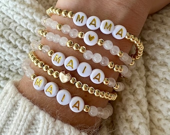 May include: A set of gold bead bracelets with white beads that spell out the names "MAIA" and "MAMA". Each bracelet has a small gold heart charm.