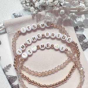 May include: Three beaded bracelets with gold beads and white beads with gold lettering. The bracelets spell out "It's Me", "I'm the Bride", and "It's Me Mi".