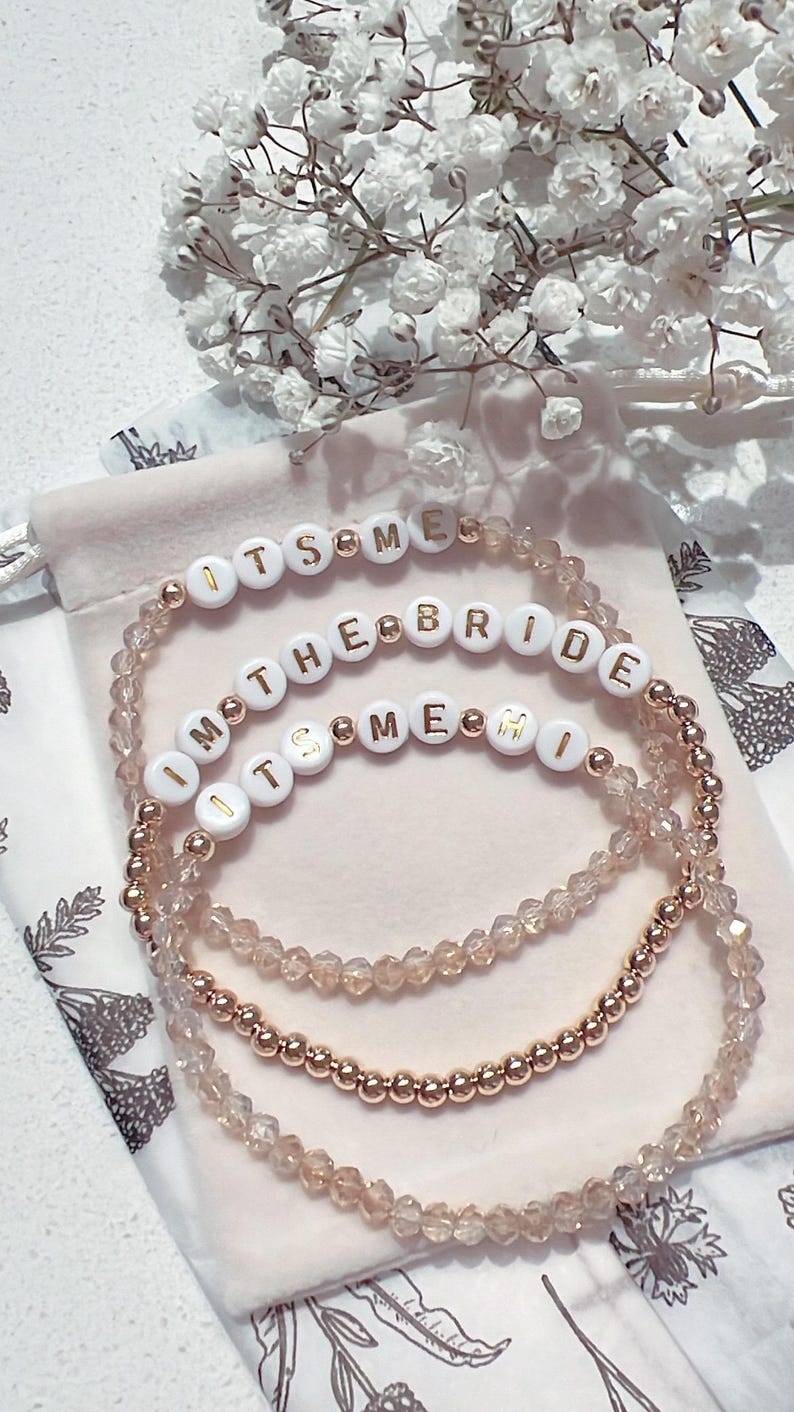May include: Three beaded bracelets with gold beads and white beads with gold lettering. The bracelets spell out "It's Me", "I'm the Bride", and "It's Me Mi".