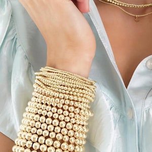 May include: A wrist displaying multiple gold beaded bracelets, with varying bead sizes creating a layered look. A delicate gold necklace with a small pendant is also visible. The background features a light blue shirt, complementing the gold jewellery.