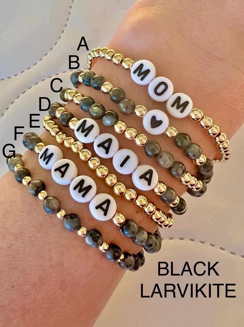 May include: Seven bracelets made with black larvikite beads and gold beads. The bracelets have white letter beads spelling out "MOM", "MAIA", and "MAMA". One bracelet has a heart bead.