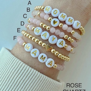 May include: Six gold beaded bracelets with white letter beads spelling out "MOMMY", "MAYA", and "MAMA". Each bracelet also has a small gold heart charm. The bracelets are on a person's wrist and are layered on top of each other. The background is a light pink colour. The text "ROSE QUARTZ" is at the bottom of the image.