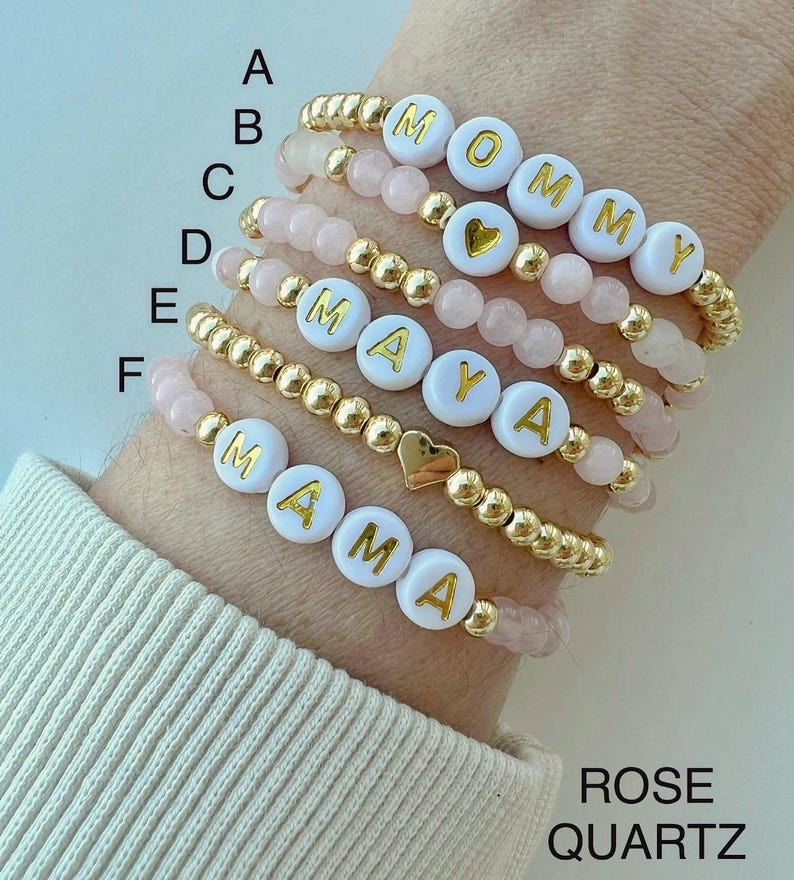 May include: Six gold beaded bracelets with white letter beads spelling out "MOMMY", "MAYA", and "MAMA". Each bracelet also has a small gold heart charm. The bracelets are on a person's wrist and are layered on top of each other. The background is a light pink color. The text "ROSE QUARTZ" is at the bottom of the image.