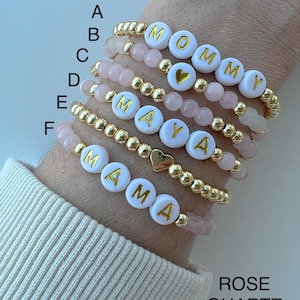 May include: Six bracelets with gold beads and white beads with gold letters spelling out "MOMMY", "MAYA", and "MAMA". The bracelets also have pink rose quartz beads.