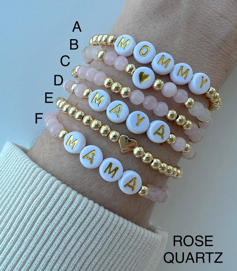 May include: Six bracelets with gold beads and white beads with gold letters spelling out "MOMMY", "MAYA", and "MAMA". The bracelets also have pink rose quartz beads.