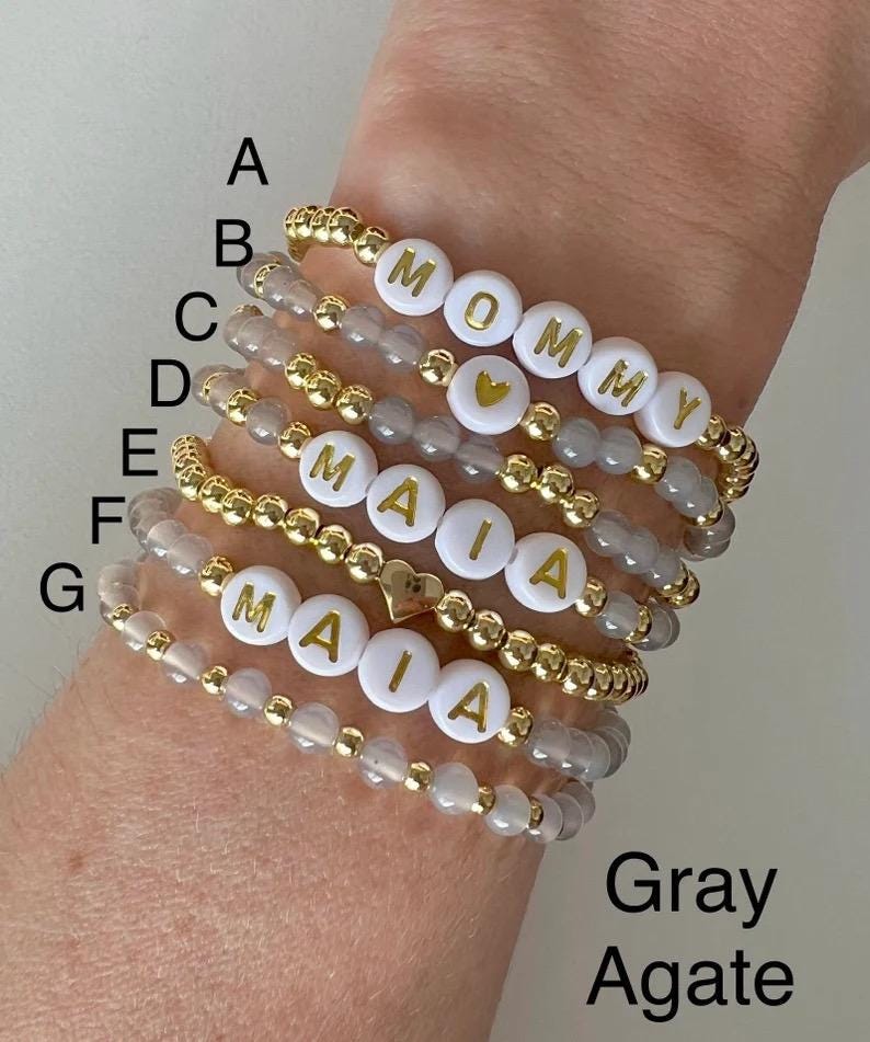 May include: Seven bracelets stacked on a wrist. The bracelets are made of gray agate beads and gold beads with white letter beads spelling out the names "Mommy" and "Maia".
