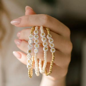 Peut inclure: Quatre bracelets en perles blanches avec des lettres dorées qui épellent "MAMMA", "NANA", "MOMMY" et "GRAMMY".
