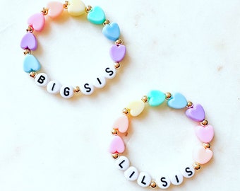 May include: Two bracelets with pastel heart beads and gold spacers. The bracelets spell out "BIG SIS" and "LIL SIS" in white letters.