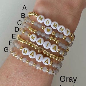 May include: Seven gray agate beaded bracelets with gold accents. Each bracelet features white beads with gold letters spelling out the names "Mommy" and "Maia".