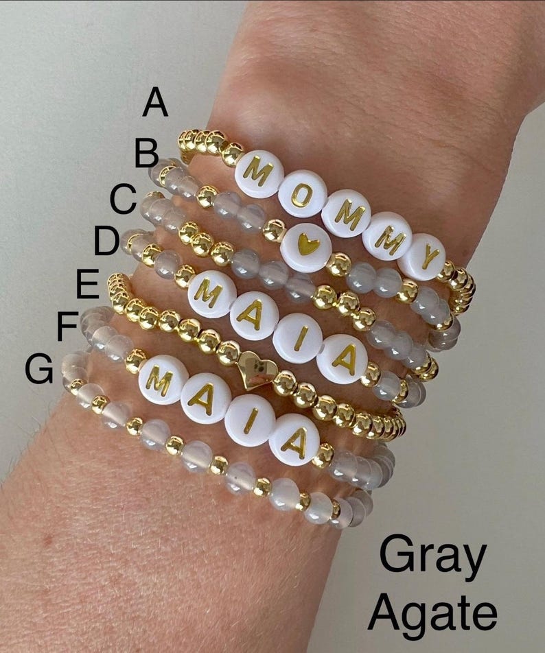 May include: Seven gray agate beaded bracelets with gold accents. Each bracelet features white beads with gold letters spelling out the names "Mommy" and "Maia".