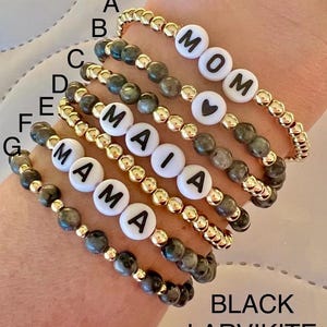 May include: Seven bracelets made with black larvikite beads and gold beads. The bracelets have white letter beads spelling out "MOM", "MAIA", and "MAMA". One bracelet has a heart bead.