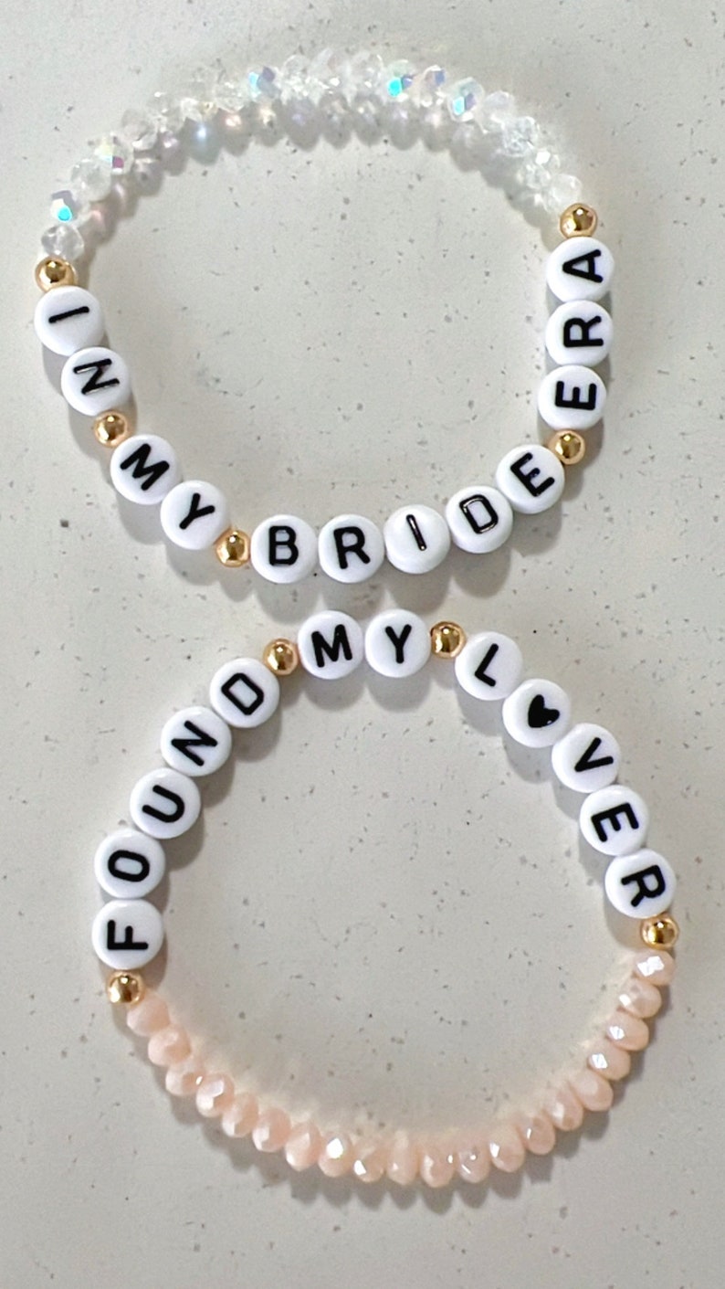 May include: Two beaded bracelets with white beads and gold spacers. The top bracelet has clear beads and spells "I'm My Bride Era". The bottom bracelet has pink beads and spells "Found My Lover".