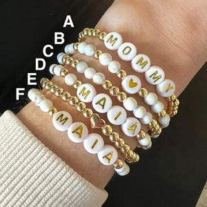 May include: A set of six bracelets made with white howlite beads and gold accents. The bracelets spell out the names "Mommy" and "Maia".