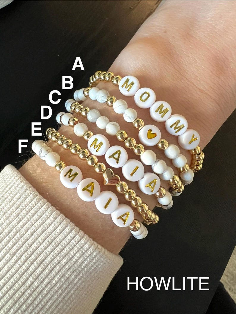 May include: A set of six bracelets made with white howlite beads and gold accents. The bracelets spell out the names "Mommy" and "Maia".