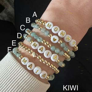 May include: A stack of six bracelets with gold beads and white beads with gold letters spelling out the names "Mommy" and "Maia". The bracelets are on a person's wrist. The bracelets are made with kiwi jasper beads.