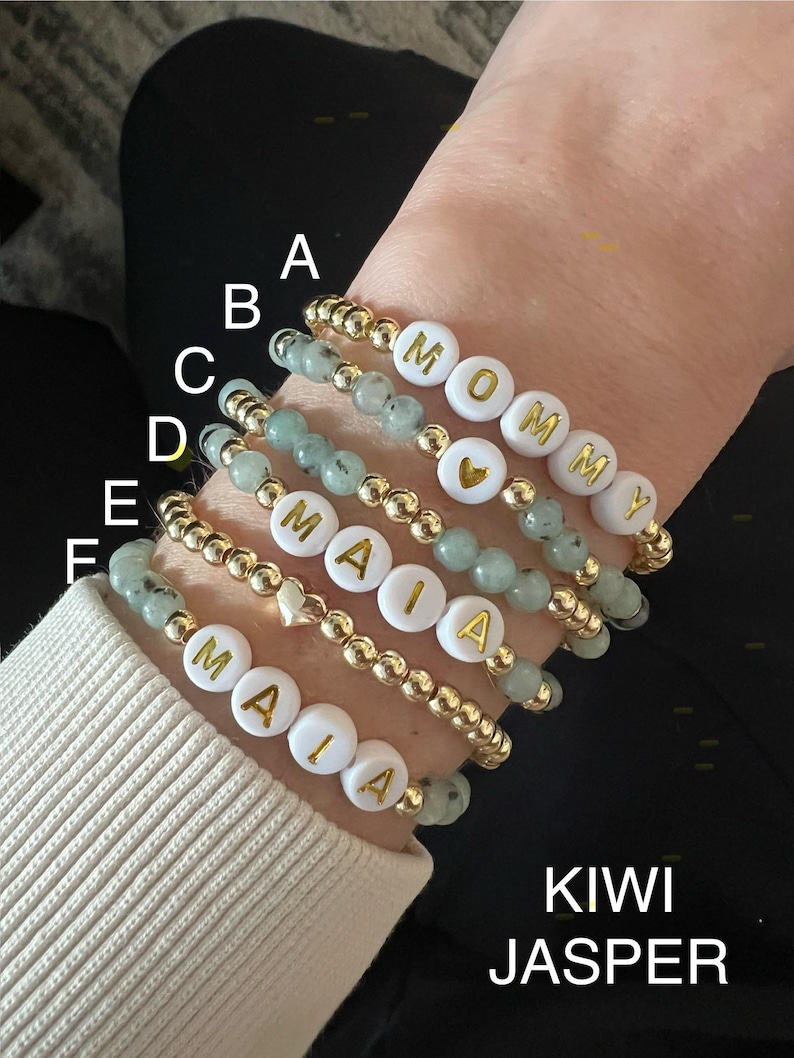 May include: A stack of six bracelets with gold beads and white beads with gold letters spelling out the names "Mommy" and "Maia". The bracelets are on a person's wrist. The bracelets are made with kiwi jasper beads.