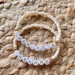 May include: Two beaded bracelets with white letter beads spelling "CHARLOTTE" and "IRELAND". The bracelets are made of off-white beads with gold accents. The background is a woven, tan surface.
