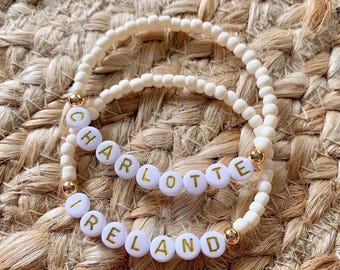 May include: Two beaded bracelets with white letter beads spelling "CHARLOTTE" and "IRELAND". The bracelets are made of off-white beads with gold accents. The background is a woven, tan surface.