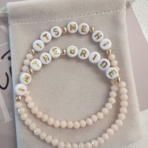 May include: Two white beaded bracelets with gold lettering spelling "It's Me, The Bride". The bracelets are on a light pink velvet background.