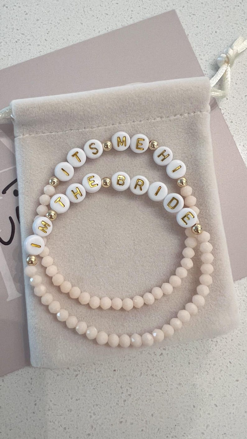 May include: Two white beaded bracelets with gold lettering spelling "It's Me, The Bride". The bracelets are on a light pink velvet background.