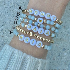 May include: Six beaded bracelets with gold beads and white beads with gold lettering. The bracelets spell out "MOMMY" and "MAIA". The beads are aquamarine.