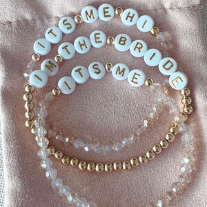 May include: Three gold beaded bracelets with white beads that spell out "It's Me", "I'm The Bride", and "It's Me".