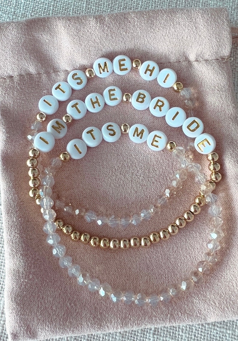 May include: Three gold beaded bracelets with white beads that spell out "It's Me", "I'm The Bride", and "It's Me".