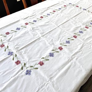 May include: A rectangular white tablecloth featuring a central embroidered border of small green vines with alternating purple and pink flowers. The embroidery forms a continuous rectangular frame pattern across the surface of the fabric.