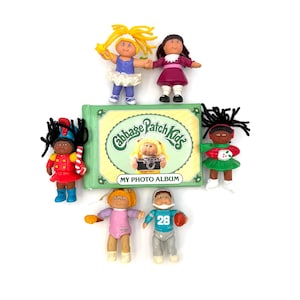 May include: A Cabbage Patch Kids My Photo Album with a green cover and a central illustration of a doll holding a camera, surrounded by six small plastic figurines including a ballerina, a girl in a maroon dress, a drummer, a holiday figure, a child in pink, and a football player.