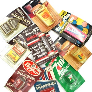 May include: Assortment of vintage pencil sharpeners featuring various brands like Hershey's Kisses, Tootsie Roll, Dr Pepper, 7 Up, and Burger King. The sharpeners are packaged in colorful cardboard boxes with brand logos and product names.