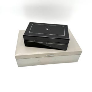May include: Two rectangular boxes, one stacked on top of the other. The top box is black with a silver trim and a small, stylized design on the lid. The bottom box is a light gray color.