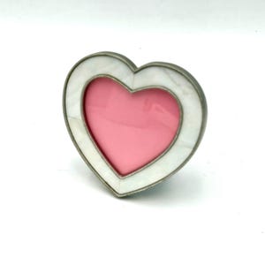 May include: A heart-shaped picture frame featuring a white, marble-like border with a metallic silver-toned edge. The center of the frame contains a solid pink insert, creating a distinct contrast between the white outer border and the pink interior.