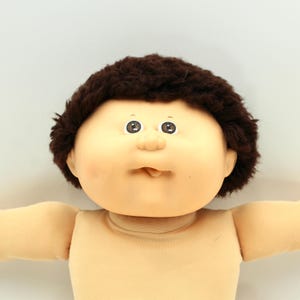 May include: A Cabbage Patch Kid doll with a tan face, brown hair, and brown eyes. The doll has a tan body and is wearing a tan shirt. The doll's arms are outstretched.