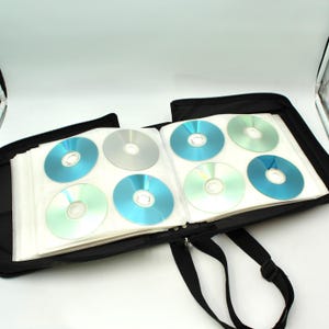 May include: Black CD carrying case, open to reveal several CDs in clear plastic sleeves. The CDs are silver, blue, and green. The case has a black shoulder strap and a zipper closure.