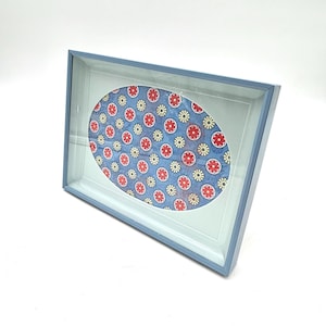 May include: A blue framed picture with an oval floral design. The design features red and yellow flowers on a blue background. The frame is light blue and has a glass cover.