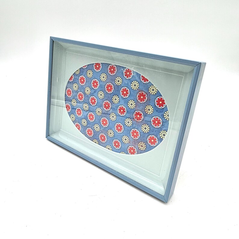 May include: A blue framed picture with an oval floral design. The design features red and yellow flowers on a blue background. The frame is light blue and has a glass cover.