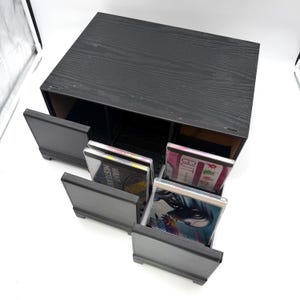May include: A black wooden storage cabinet with a visible wood grain texture, featuring two pull-out drawers. The drawers contain several compact disc cases with visible cover art, including one labeled with the text "WANT MISTEAK".