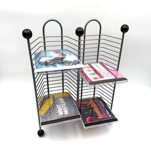 May include: A gray metal wire rack with two vertical sections for storing media cases. The rack features black spherical finials at the top and bottom corners. Four plastic cases are inserted into the wire slots, displaying various graphic covers.
