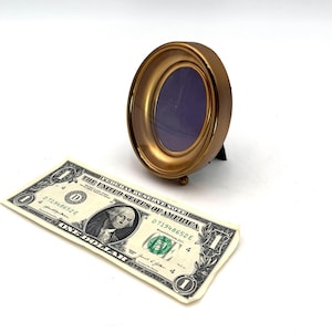 May include: A small, oval-shaped, gold-colored picture frame with a dark blue interior. The frame is supported by a small black stand. A one-dollar bill is in the foreground.