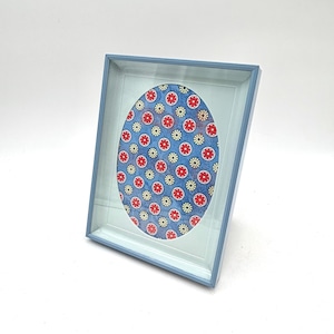 May include: A blue picture frame with a light blue interior. The frame holds an oval-shaped image with a blue background and a pattern of red and yellow flowers. The frame is angled slightly, showcasing its design.