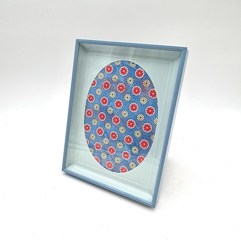 May include: A blue picture frame with a light blue interior. The frame holds an oval image with a blue background and a pattern of red and yellow flowers. The frame is angled, displaying its design.