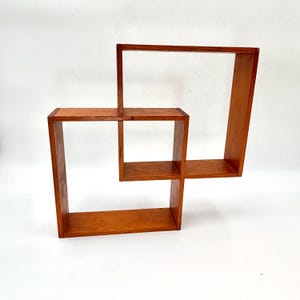 May include: A wall-mounted wooden shelf unit featuring an interconnected geometric design. The structure consists of two rectangular frames joined together, creating multiple open compartments with a warm, natural brown wood grain finish.